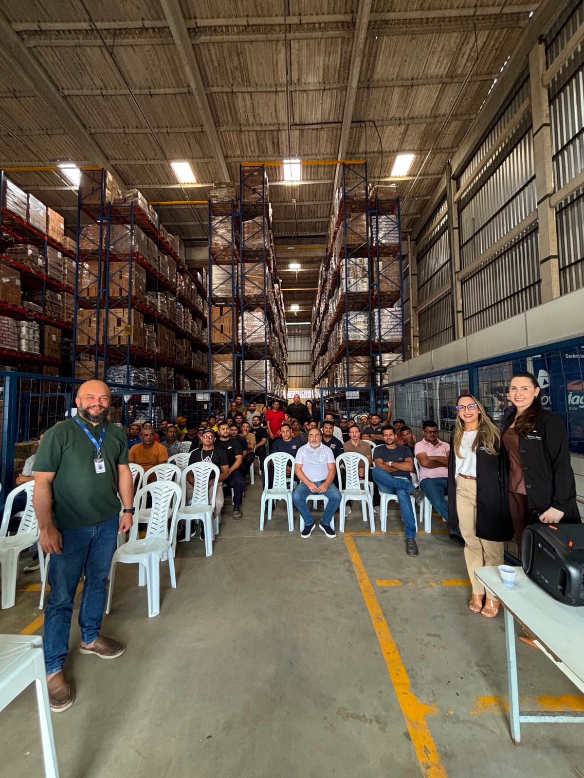 Equipe de uma distribuidora de alimentos durante o treinamento, sentada no galpão entre as prateleiras de estoque