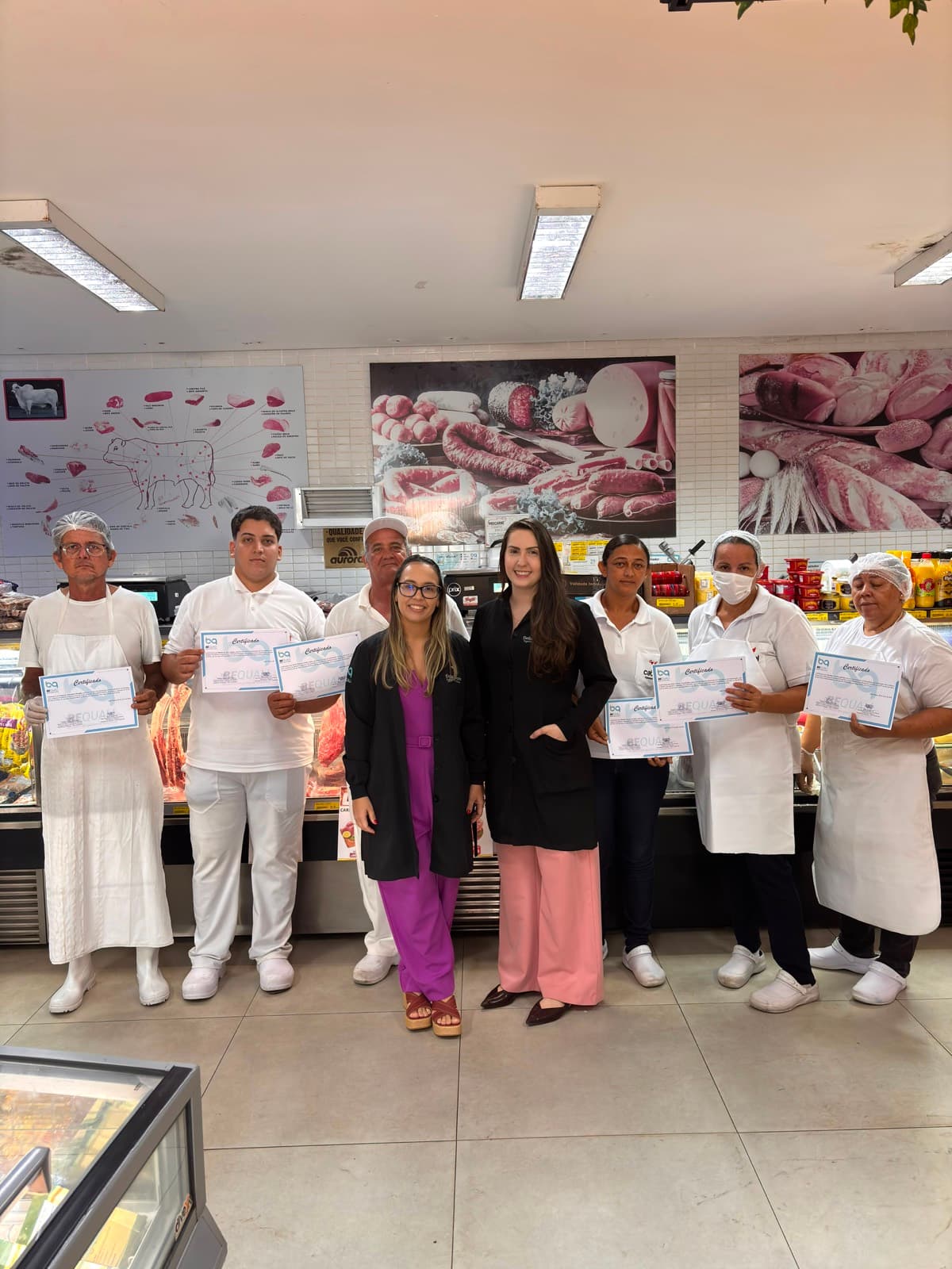 Equipe do açougue e do setor de frios de um supermercado com os certificados de treinamento, em frente ao balcão refrigerado, ao lado das consultoras da BeQuali